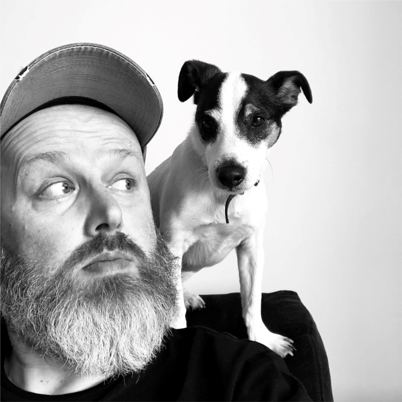 Tom Youll is a bearded person. He is wearing a baseball cap. On his shoulder sits a black and white dog.